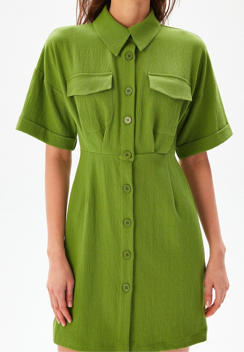 Femme portant une robe-chemise boutonnée à manches courtes de couleur vert olive texturée avec deux poches à rabat sur la poitrine et un col.