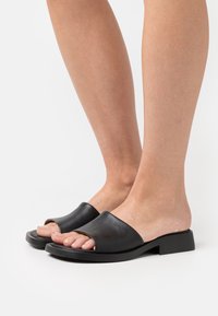 Feet wearing black open-toe slide sandals with low block heels against a plain white background.