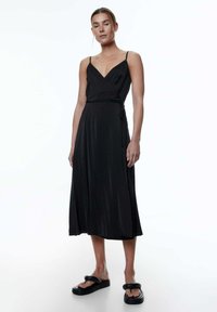 Schwarzes Midi-Kleid mit Spaghetti-Trägern, V-Ausschnitt, Wickeldesign und glattem Stoff. Kombiniert mit klobigen schwarzen Slipper-Sandalen.
