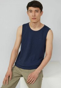 Navy blue sleeveless shirt with a round neckline, made of lightweight fabric. Worn with beige pants, featuring a subtle logo detail.