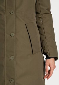 Olivgrön vattentät jacka med stora knappar, en sidoficka och en kardborreband vid ärmen. Slät textur och knälång design.