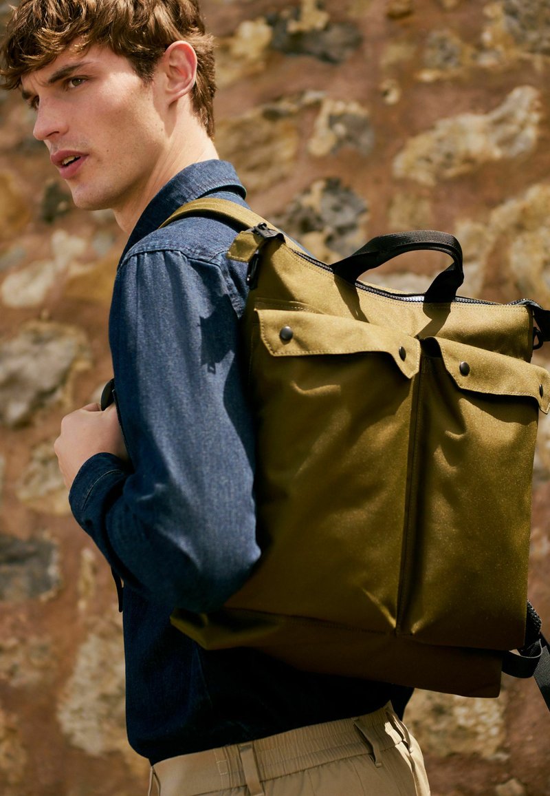 Olivgrüner Rucksack aus robustem Material mit rechteckiger Form, zwei Klappentaschen und einem oberen Griff. Getragen über einem Jeanshemd.