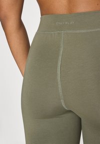 Leggings de color verde oliva con diseño sin costuras, cinturilla elástica y costuras de contraste. El material es suave y tiene una forma ajustada.