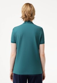 Person med kort lyst hår iført en teal kortærmet polo-shirt og mørke bukser, vendt væk fra en ensfarvet hvid baggrund.