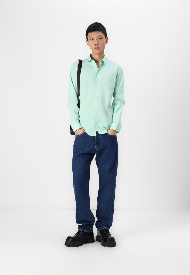 Camisa de manga larga verde menta, de tela ligera, con botones en la parte frontal; combinada con jeans de mezclilla azul holgados y zapatos negros gruesos; bolso de hombro.