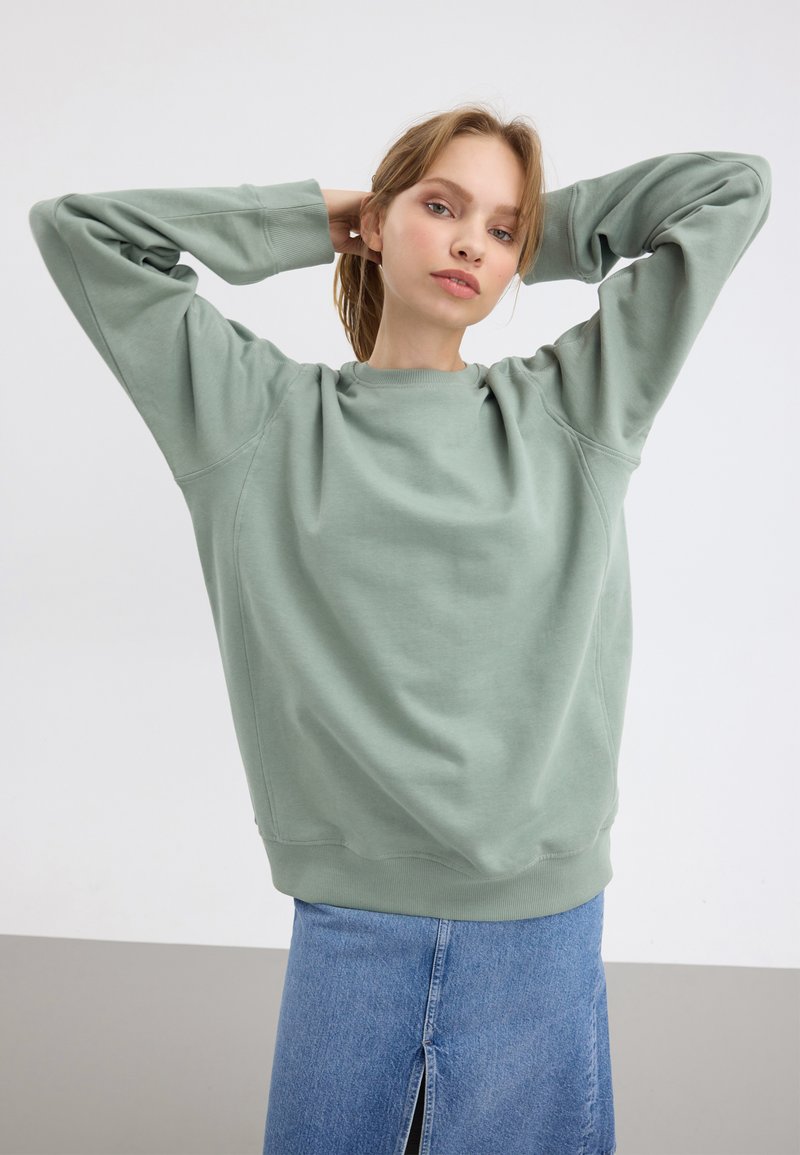 Sweatshirt vert menthe avec une coupe décontractée, des manches raglan et des poignets côtelés. Porté avec une jupe en denim avec une fente à l'avant.