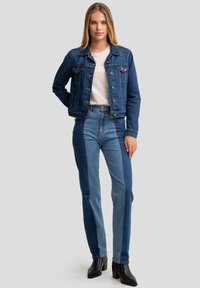 Denimjacket in een donkerblauwe was met knoopsluiting, gecombineerd met hoog- taille spijkerbroek met kleurblokken in verschillende tinten blauw, zwarte haklaarzen.
