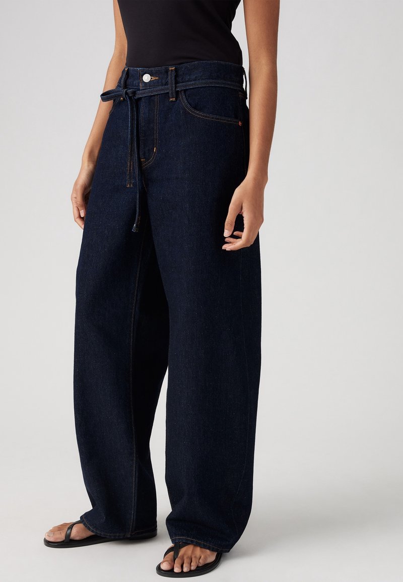 Femme portant un jean large foncé avec une ceinture en tissu nouée à la taille, associée à des tongs noires et un haut noir sans manches.