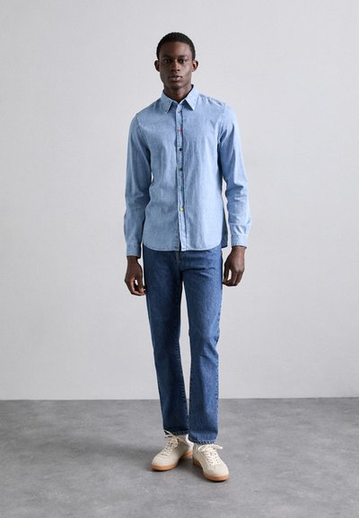 Camisa de mezclilla de botones, de manga larga y color azul claro. Combinada con jeans azul oscuro y zapatillas beige, de pie contra un fondo neutro.