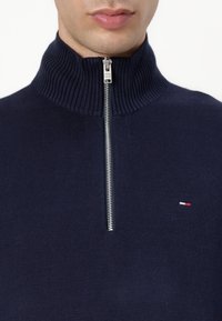 Navyblauwe gebreide trui met een hoge geribbelde kraag en zilveren rits. Voorzien van een klein geborduurd logo in rood, wit en blauwe kleuren op de borst.