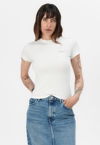Balta trumpa rankovėmis marškinėliai, pagaminti iš medvilnės, su mažu "Tommy Jeans" logotipu ant krūtinės, derinami su mėlynu džinsiniu sijonu.