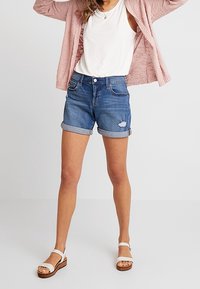 Džínové šortky s úzkým střihem, ohrnutými lemy a vyšisovanými detaily. V kombinaci s bílým tílkem a růžovým pleteným svetrem. Hnědé platformové sandály.