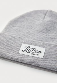 Gorro de lana gris hecho de material suave, con una etiqueta blanca que dice "Lil' Boo Copenhagen" en texto cursivo negro. Diseño simple y versátil.