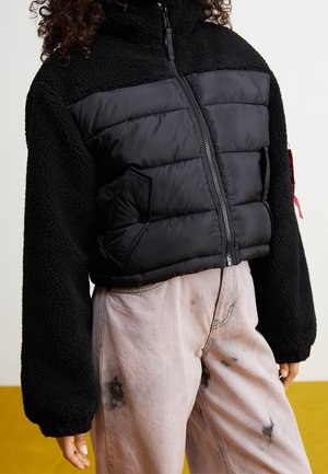 Person trägt eine schwarze wattierte und Fleece-Reißverschlussjacke mit hellrosa Hose, die dunkle Flecken hat.