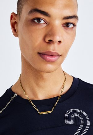 Collana in oro con pendente rettangolare allungato, indossata sopra una camicia blu navy con dettaglio a zip e design a cucitura bianca.