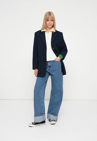 Blazer blu navy con chiusura a bottone singolo, abbinato a una polo bianca e jeans blu larghi. Il modello indossa sneakers nere con suole bianche.