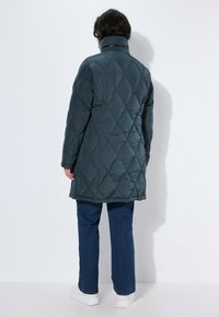 Cappotto imbottito lungo in turchese scuro con colletto alto, caratterizzato da un motivo a diamante e una texture liscia. Abbinato a jeans blu e sneakers bianche.