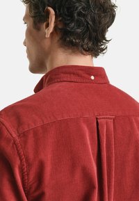 Chemise en velours côtelé rouge avec un col rabattu, un pli au dos et un petit bouton blanc au niveau du cou. Le tissu présente un motif côtelé texturé.