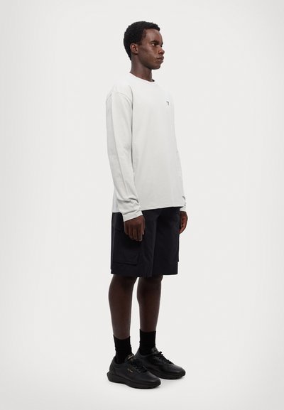 Lange mouw lichtgrijze shirt gecombineerd met zwarte shorts. Gemaakt van een gestructureerde stof met een subtiel logo op het shirt. Zwarte sneakers maken de outfit compleet.