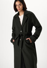 Σκούρο πράσινο παλτό με ζώνη, κατασκευασμένο από υφασμα με υφή, διαθέτοντας μεγάλο γιακά, κουμπιά μπροστά και πλαϊνές τσέπες. Απλός και λειτουργικός σχεδιασμός.