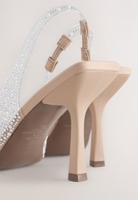 Sandales à talons hauts beige avec des brides transparentes décorées de petits strass, vues de dos sur un fond clair uni.