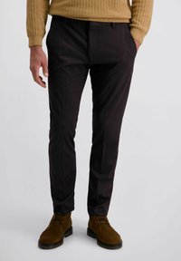 Mann in dunklen, schmal geschnittenen Hosen, braunen Wildleder-Schnürschuhen und einem beigen gerippten Pullover mit den Händen in den Taschen.