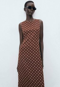 Brown sleeveless dress featuring cream polka dots, smooth texture, and a straight silhouette. Styled with black sunglasses.