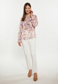 Bluse mit Blumenmuster in hellbeige, mit Rüschen und Bändern am Ausschnitt, kombiniert mit weißen, taillierten Hosen und braunen Wildlederhighheels.