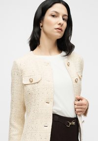 Sötét vállig érő hajú nő, krémszínű tweed dzsekit visel gyöngy gombokkal, fehér felsővel és sötét övvel megkötött nadrággal.