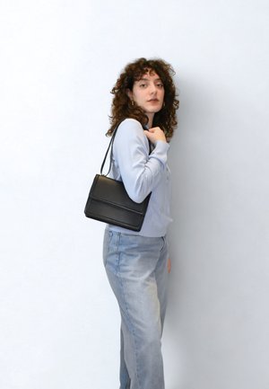 Czarna skórzana torba na ramię o prostokątnym kształcie, z przednią klapką i cienkim paskiem na ramię, pokazana na osobie noszącej jasnoniebieską bluzkę i dżinsy.