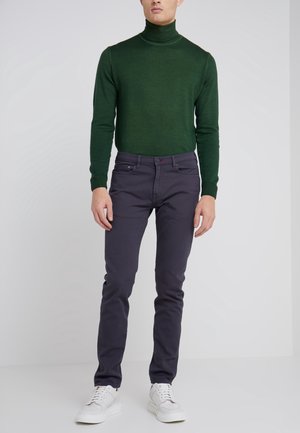 Mann trägt einen dunkelgrünen Rollkragenpullover, dunkle schlanke Hosen und weiße Turnschuhe und steht vor einem schlichten hellen Hintergrund.