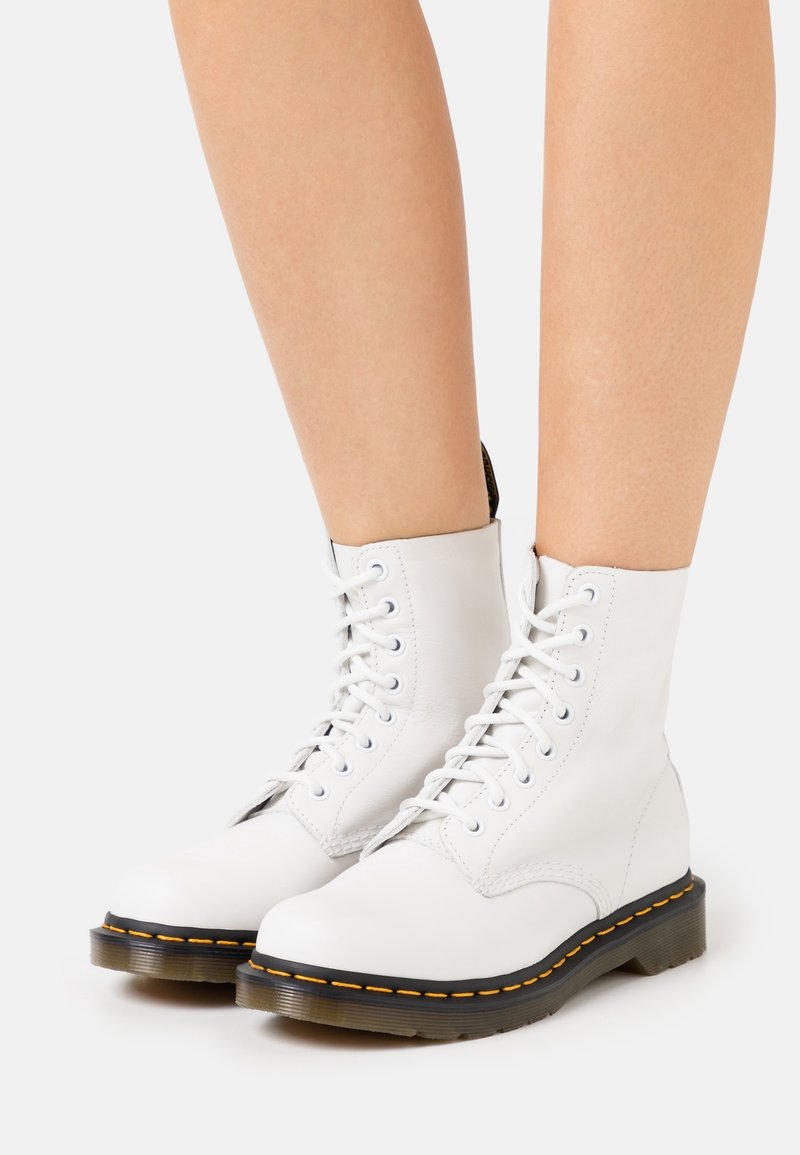 Bottines blanches à lacets avec semelles épaisses noires et surpiqûres jaunes portées sur des jambes nues sur un fond uni.