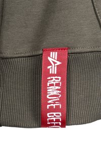 Felpa in cotone verde oliva con orlo a costine. Presenta un'etichetta in tessuto rossa con testo bianco che reca la scritta "RIMUOVERE PRIMA".