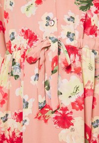 Detailní pohled na růžovou květinovou látku šatů s červenými, bílými a zelenými květy, zavázaným páskem a růžovým knoflíkem nad pasem.