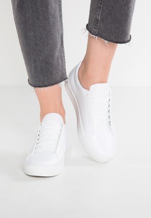 Personne portant des baskets en cuir blanc et un jean gris raccourci avec des bords effilochés, debout sur un sol blanc.