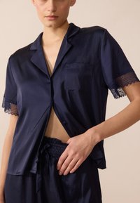 Jūras spēku satīna pidžamas komplekts ar īsām piedurknēm, dekorēts ar mežģīnēm un ar pogu aizdari, kā arī biksēm ar elastīgu jostas vietu.