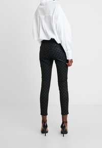 Persona vistiendo pantalones ajustados negros con lunares, una camisa blanca oversized y sandalias de tacón alto negras, de pie contra un fondo liso.