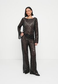 Tenue deux-pièces noire à sequins comprenant un haut à manches longues avec poignets évasés et un pantalon ajusté évasé, reflétant la lumière avec une texture brillante.