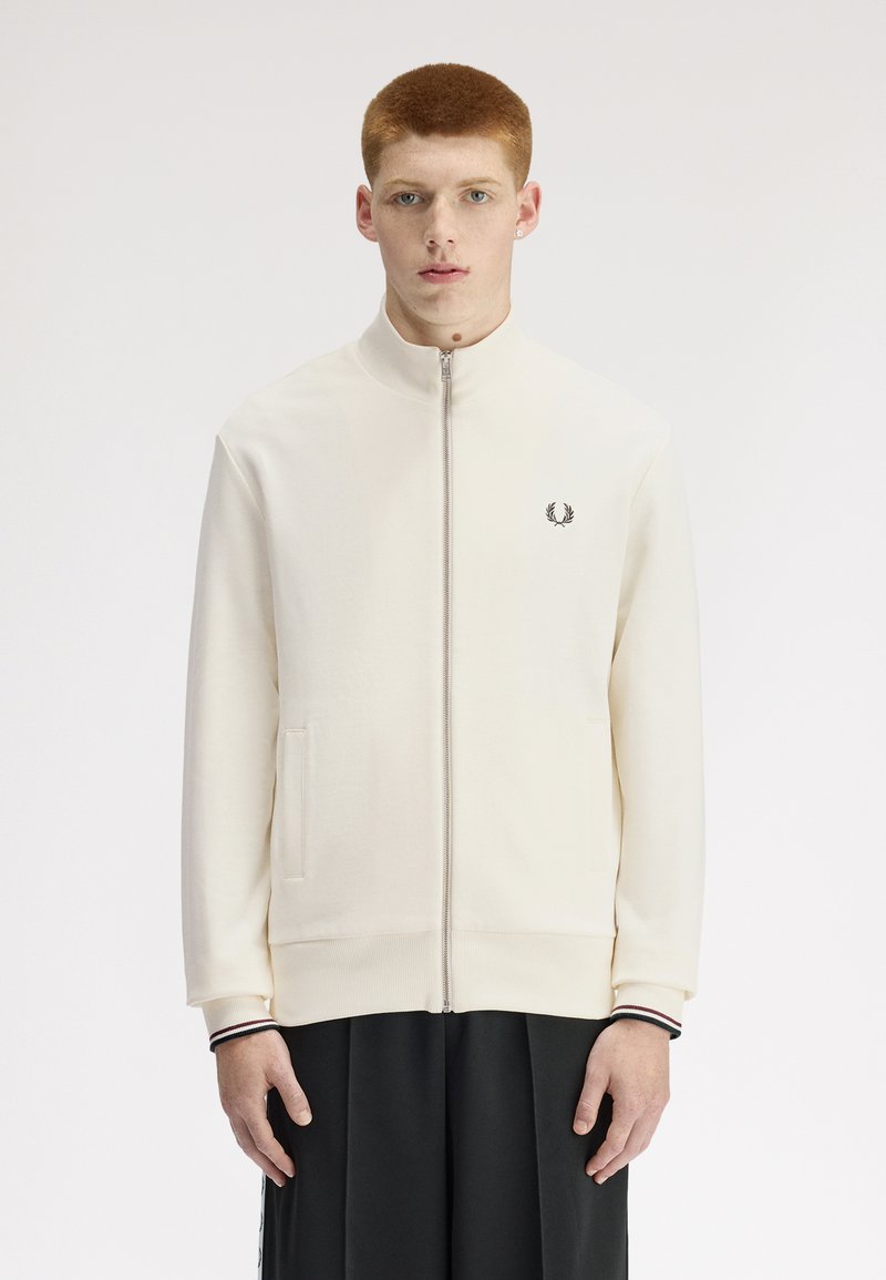 Jeune homme aux cheveux roux courts portant une veste crème zippée avec des poches latérales et un pantalon noir, debout devant un fond uni.