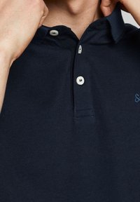 Mörkblå pikétröja i texturerad bomull, med klassisk krage, två vita knappar och en liten broderad logotyp på vänster bröst.