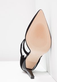 Chaussure à talon haut en daim noir pointue reposant sur le côté, montrant la semelle beige lisse avec une zone de grip à motifs et un talon élancé.