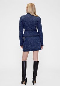 Komplet mornarsko moder tweed blazer in krilo z teksturirano površino, z pasom v pasu in kontrastnimi denim poudarki, v kombinaciji s črnimi škornji do kolen.