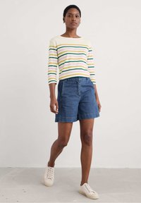 Striped long-sleeve top in yellow, green, and pink; paired with dark blue denim shorts. Model wearing beige sneakers, standing on a light floor.