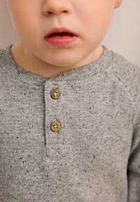 Szürke bordázott hosszúujjú ing, apró pöttyökkel, három barna gombbal a nyakán és egyszerű, klasszikus kialakítással.
