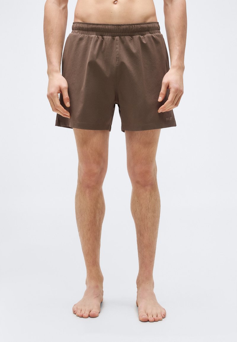 Άτομο με γυμνό στήθος, όρθιο και ξυπόλητο, φοράει καφέ σορτς με ελαστική μέση, σε απλό ανοιχτό φόντο.