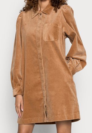 Frau trägt ein langärmeliges Cordkleid in Kamelbraun mit Brusttasche und durchgehender Knopfleiste.