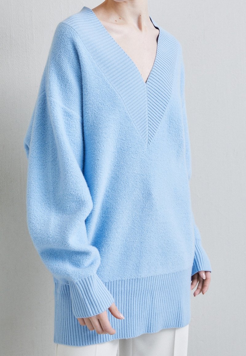 Personne portant un pull bleu clair ample avec un col en V profond et de longues manches et ourlets côtelés, associé à un pantalon blanc.