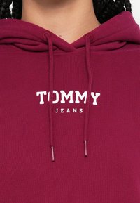 Gros plan d’une personne portant un sweat à capuche couleur bordeaux avec "TOMMY JEANS" imprimé en blanc sur la poitrine et des cheveux tressés visibles.
