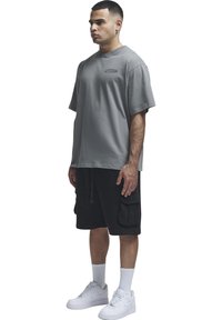 Homme debout en t-shirt gris oversize, short cargo noir, chaussettes blanches hautes et baskets blanches sur fond blanc.