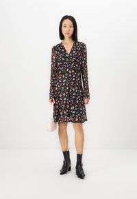 Robe noire à fleurs avec des manches longues, un décolleté en V et un détail avant torsadé ; ornée de fleurs roses et violettes, associée à des bottines noires et à un sac rose.